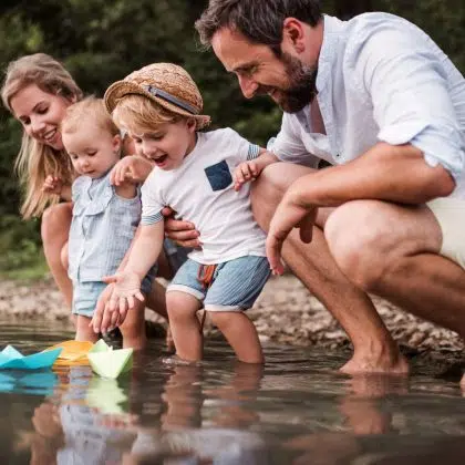 A young family with two toddler children outdoors by the river in summer, playing with paper boats.