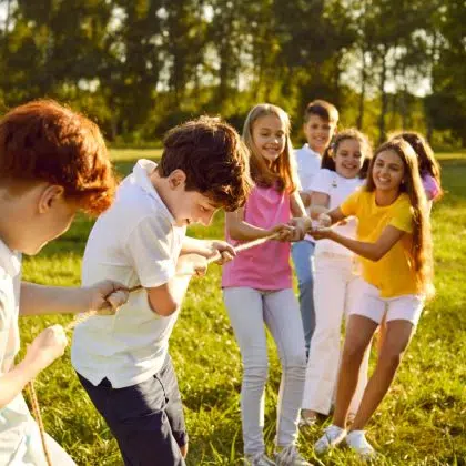 Group of a happy joyful smiling children playing together in tug-of-war with a rope in the park on summer holidays. Cheerful school kids having fun on green grass outdoors in the camp.